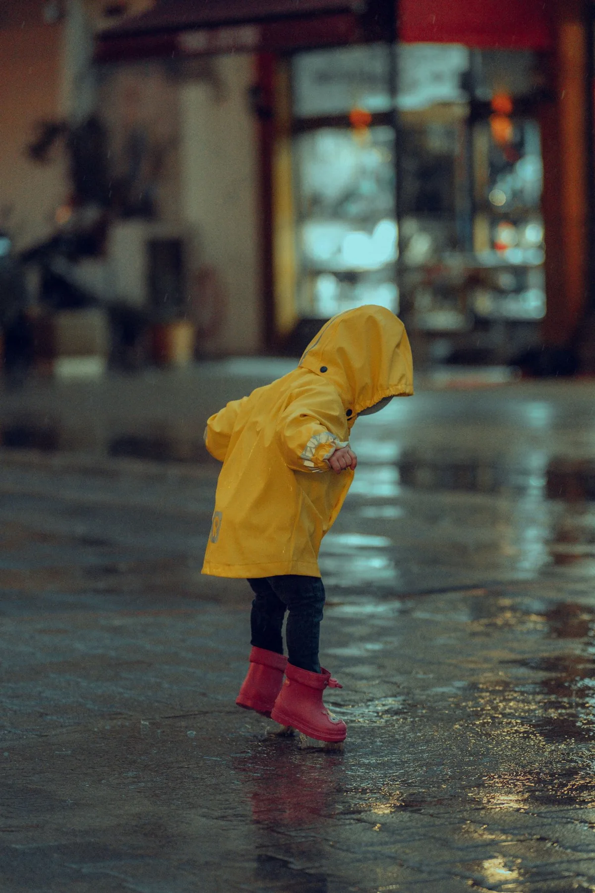 A child in a yellow coat and red boots beside a puddle
