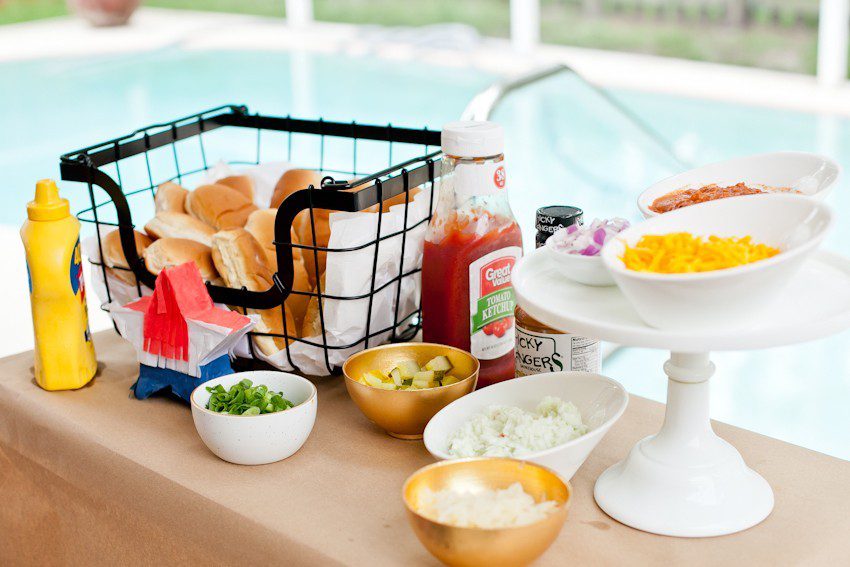  Hot Dog Bar by popular Florida lifestyle blog, Fresh Mommy Blog: image of hot dog buns in a black wire basket next to a ketchup bottle, mustard bottle, and serving bowls filled with green onions, chopped white onions, shredded cheese, and avocado slices. 
