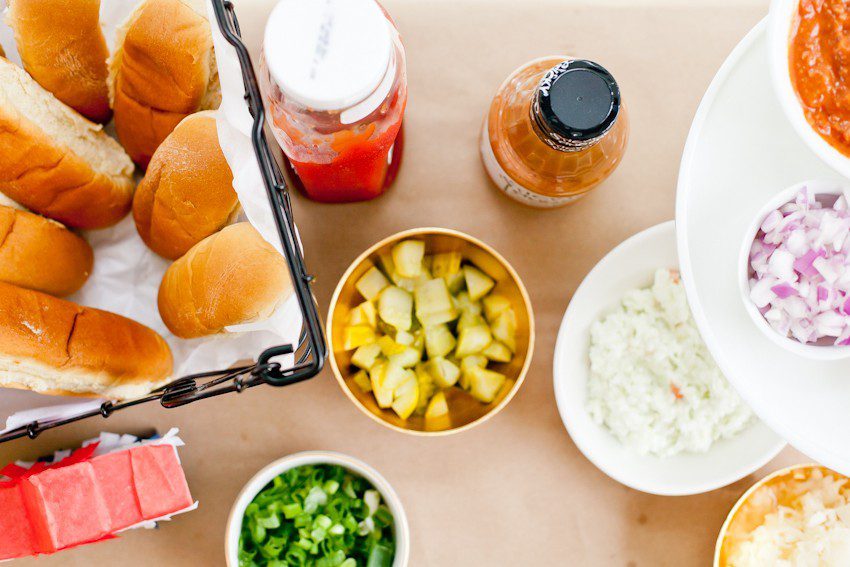 Red White and Blue fun, and a DIY Americana Hot Dog Bar! Set up toppings and flavors for all kinds of hot dogs from around the U.S. Chicago style, Boston, Detroit, Philly, Carolina hot dog and more! |  Hot Dog Bar by popular Florida lifestyle blog, Fresh Mommy Blog: image of hot dog buns in a black wire basket next to a ketchup bottle, diced white onions, diced purple onions, sliced avocados, and green onions.  