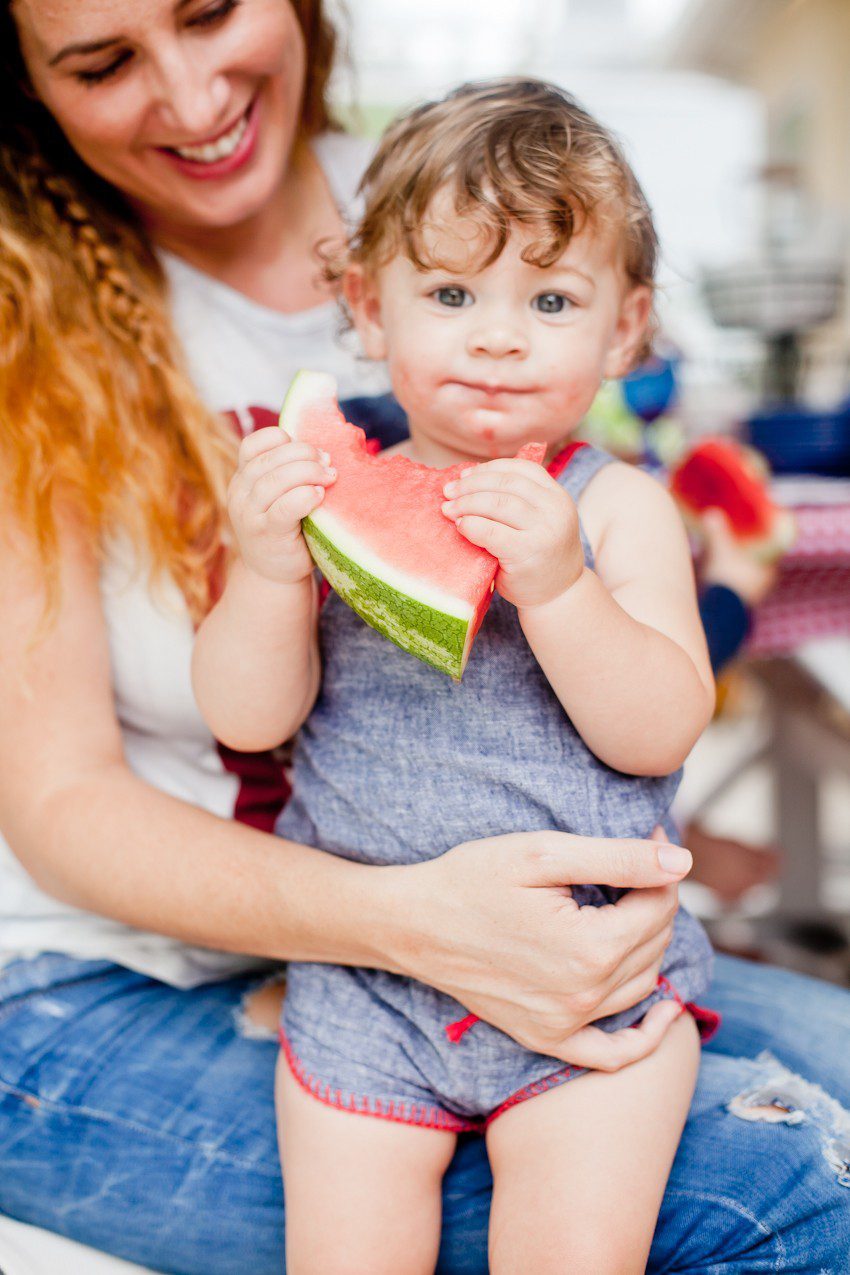 Red White and Blue fun, and a DIY Americana Hot Dog Bar! Set up toppings and flavors for hot dogs recipes from around the U.S. Chicago style, Boston, Detroit, Philly, Carolina hot dog and more! |  Hot Dog Bar by popular Florida lifestyle blog, Fresh Mommy Blog: image of a mom sitting outside and holding her baby as they eat a slice of watermelon. 