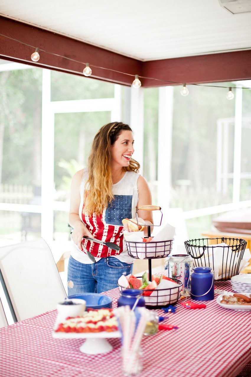 Red White and Blue fun, and a DIY Americana Hot Dog Bar! Set up toppings and flavors for all kinds of hot dogs from around the U.S. Chicago style, Boston, Detroit, Philly, Carolina hot dog and more! | Hot Dog Bar by popular Florida lifestyle blog, Fresh Mommy Blog: image of a woman wearing an American flag tank top and assembling a hot dog at a table with a red and white check table cloth, tiered black wire serving dish with watermelon slices, white serving plate of hot dogs, and American flag fruit platter. 