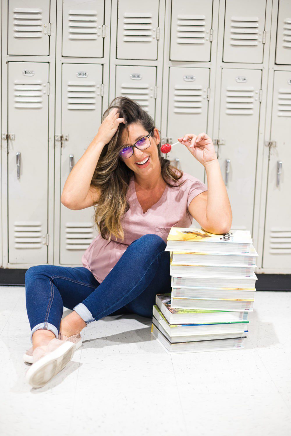 Back to School Eye Exam: How to Prepare your Child for a Successful Academic Year | Back to School Eye Exam: How to Prepare your Child for a Successful Academic Year by popular Florida lifestyle blog, Fresh Mommy Blog: image of a woman sitting in front of some school lockers wearing a pair of blue light glasses, holding a sucker, and resting her elbow on a stack of books. 