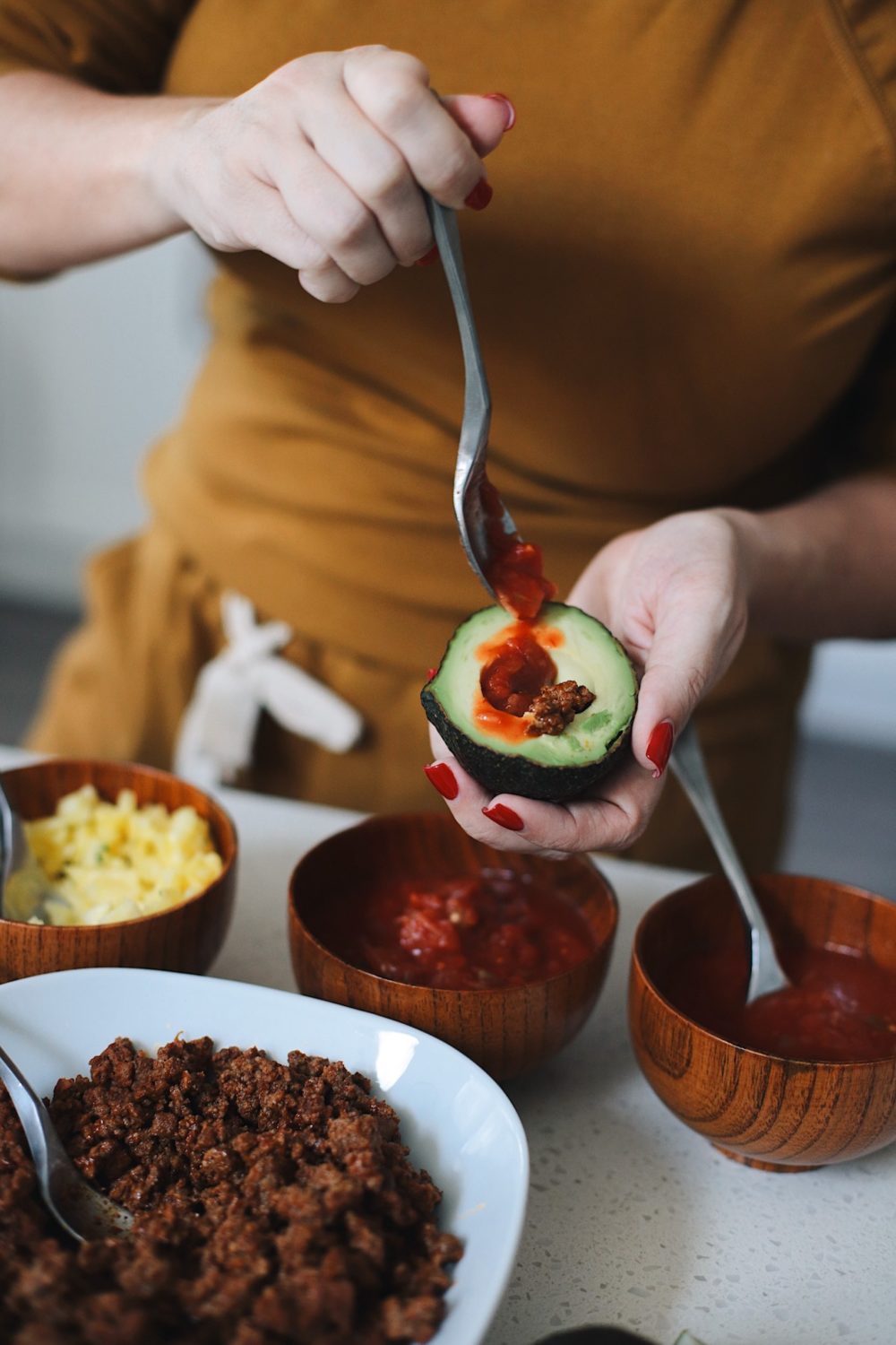 Easy Entertaining with Avocado Keto Taco Bowls recipe featured by top Florida lifestyle blog, Fresh Mommy Blog: Make-Your-Own Avo Taco Bowls from popular lifestyle blogger Tabitha Blue of Fresh Mommy Blog | Keto Taco Bowls by popular Florida lifestyle blog, Fresh Mommy Blog: image of a woman putting a spoonful of salsa in an avocado half.