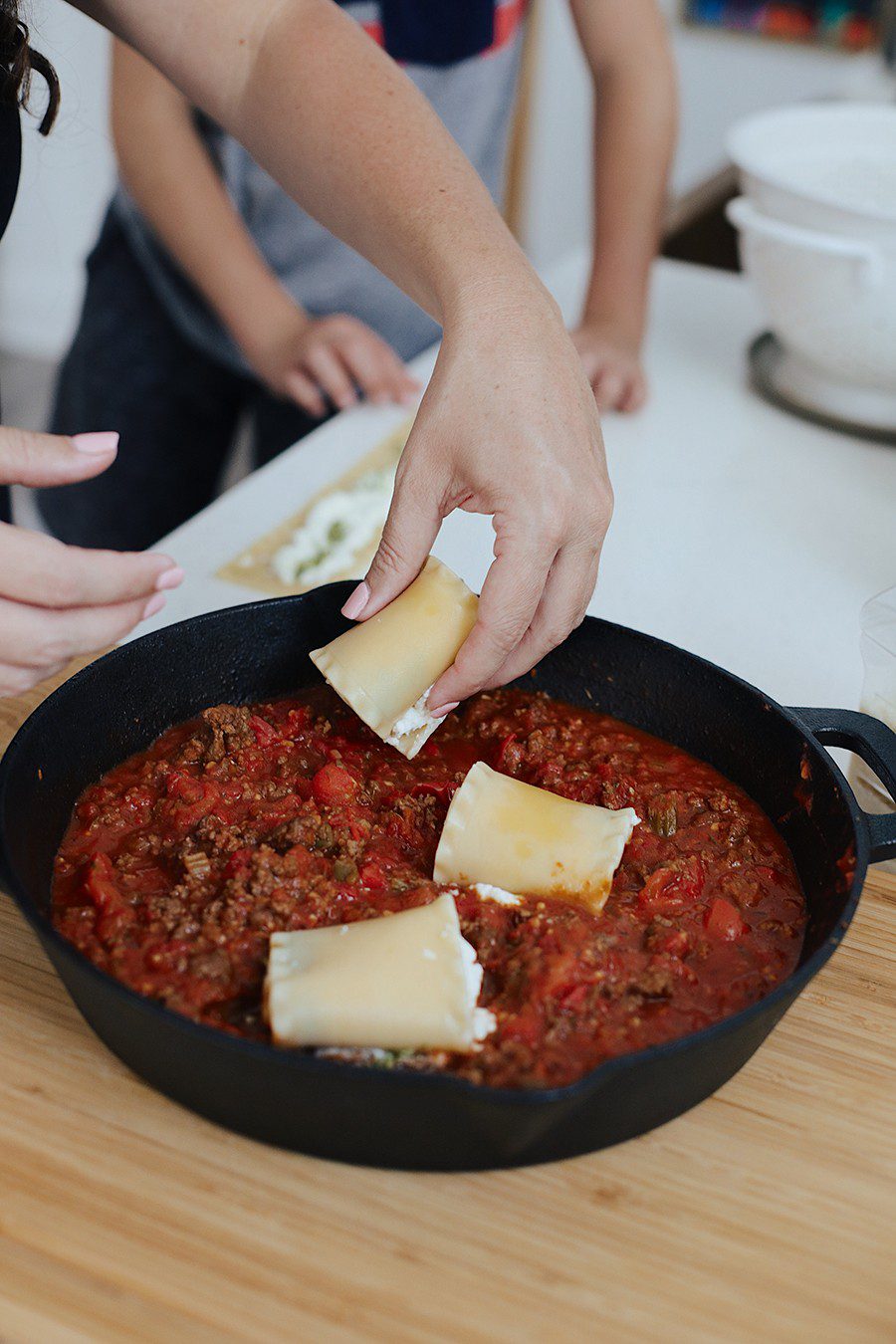 The Ultimate Cast Iron Lasagna with this lodge cast iron recipe from top Florida lifestyle blogger Tabitha Blue of Fresh Mommy Blog! Cast iron skillet cooking with kids to get family dinner on the table in 30 minutes! | Cast Iron Lasagna by popular Florida lifestyle blog, Fresh Mommy Blog: image of a woman placing rolled up lasagna noodles with pesto and ricotta cheese inside a cast iron skillet containing Bolognese sauce.  