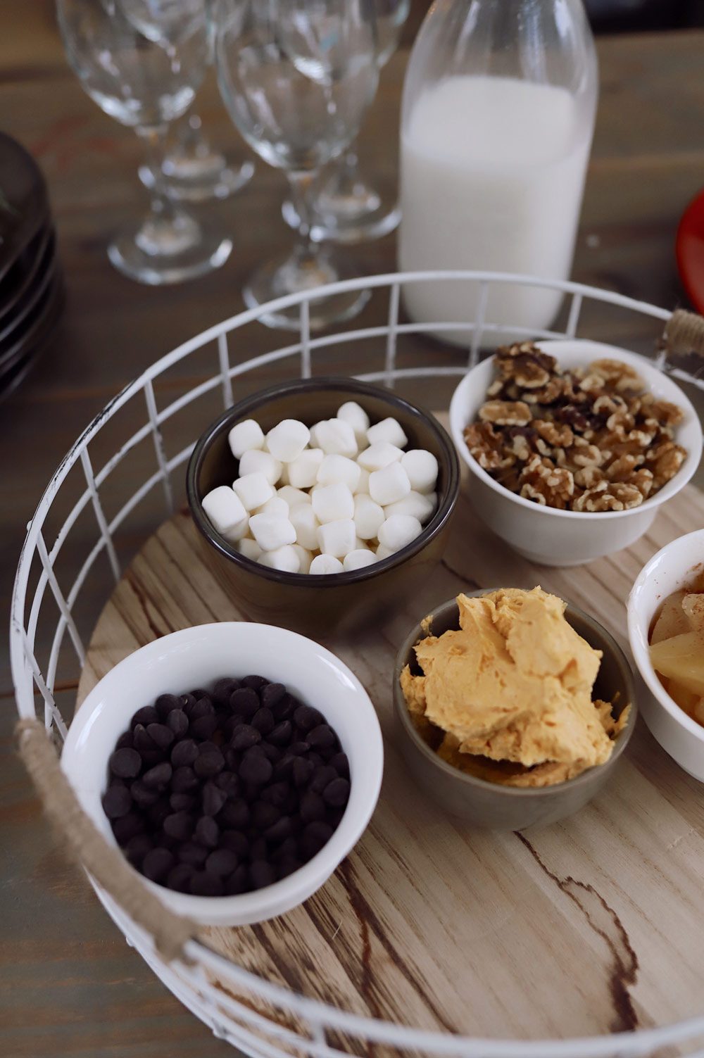 Create a festive build-your-own pancake bar with a holiday theme! Great for a Christmas brunch, Santa breakfast or delicious holiday family breakfast anytime. Plus, lots of festive pancake toppings and ideas for the best holiday pancakes! |Pancake Bar Ideas by popular Florida lifestyle blog, Fresh Mommy Blog: image of a round white wire and wood serving tray holding tiny ceramic bowls filled with mini chocolate chips, mini marshmallows, whipped gingerbread butter, walnuts, and apple slices. 