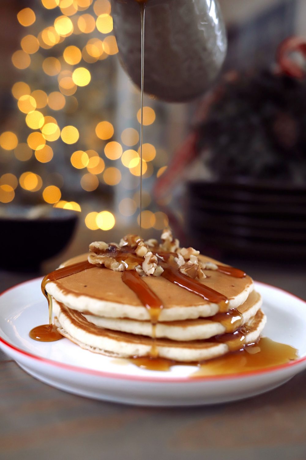 Create a festive build-your-own pancake bar with a holiday theme! Great for a Christmas brunch, Santa breakfast or delicious holiday family breakfast anytime. Plus, lots of festive pancake toppings and ideas for the best holiday pancakes! |Pancake Bar Ideas by popular Florida lifestyle blog, Fresh Mommy Blog: image of some  maple syrup being poured onto a stack of pancakes topped with walnuts. 