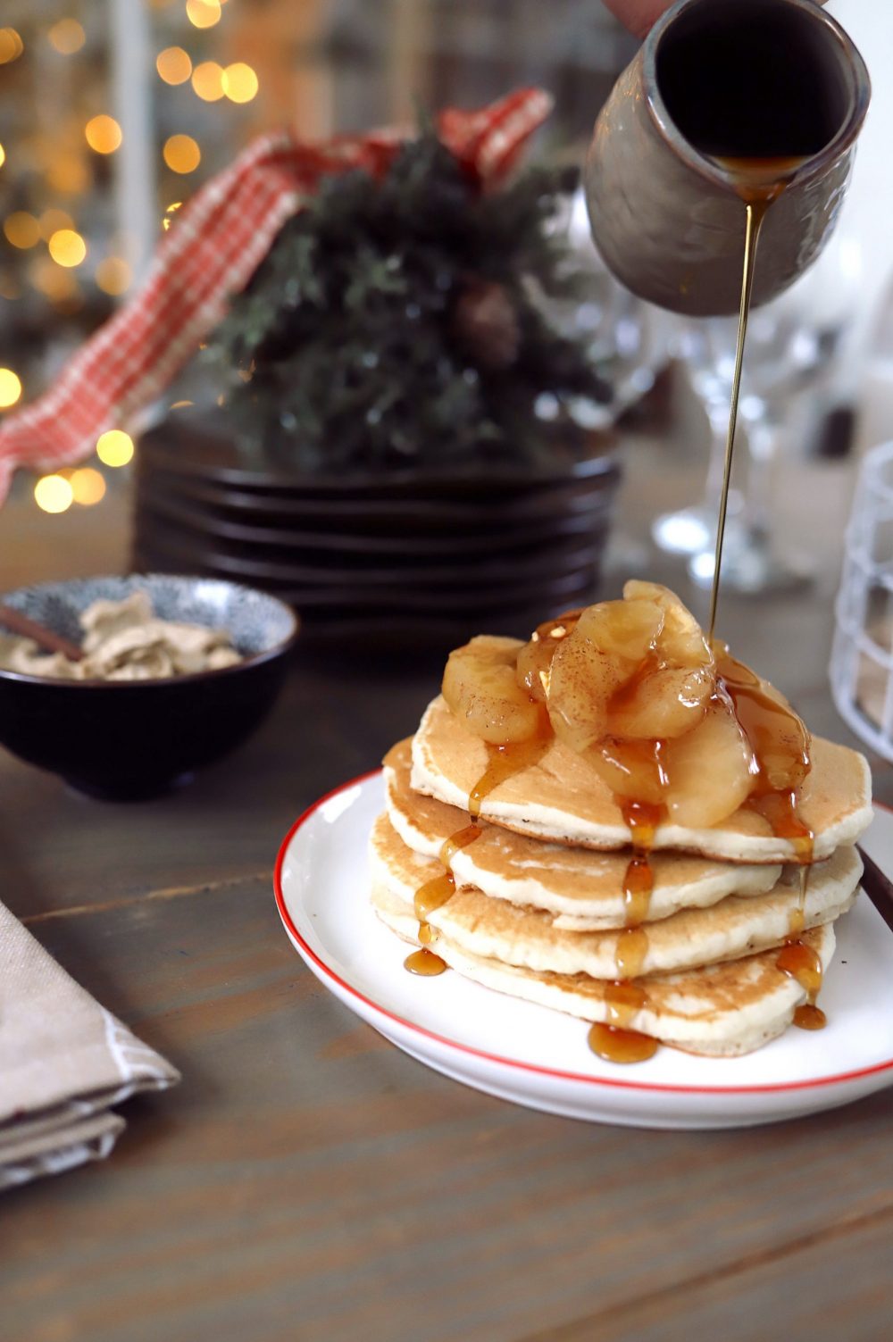 Create a festive build-your-own pancake bar with a holiday theme! Great for a Christmas brunch, Santa breakfast or delicious holiday family breakfast anytime. Plus, lots of festive pancake toppings and ideas for the best holiday pancakes! |Pancake Bar Ideas by popular Florida lifestyle blog, Fresh Mommy Blog: image of maple syrup being poured onto a stack of pancakes topped with apple slices. 