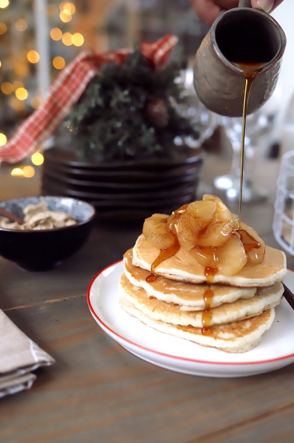 Create a festive build-your-own pancake bar with a holiday theme! Great for a Christmas brunch, Santa breakfast or delicious holiday family breakfast anytime. Plus, lots of festive pancake toppings and ideas for the best holiday pancakes! |Pancake Bar Ideas by popular Florida lifestyle blog, Fresh Mommy Blog: image of maple syrup being poured onto a stack of pancakes topped with apple slices.